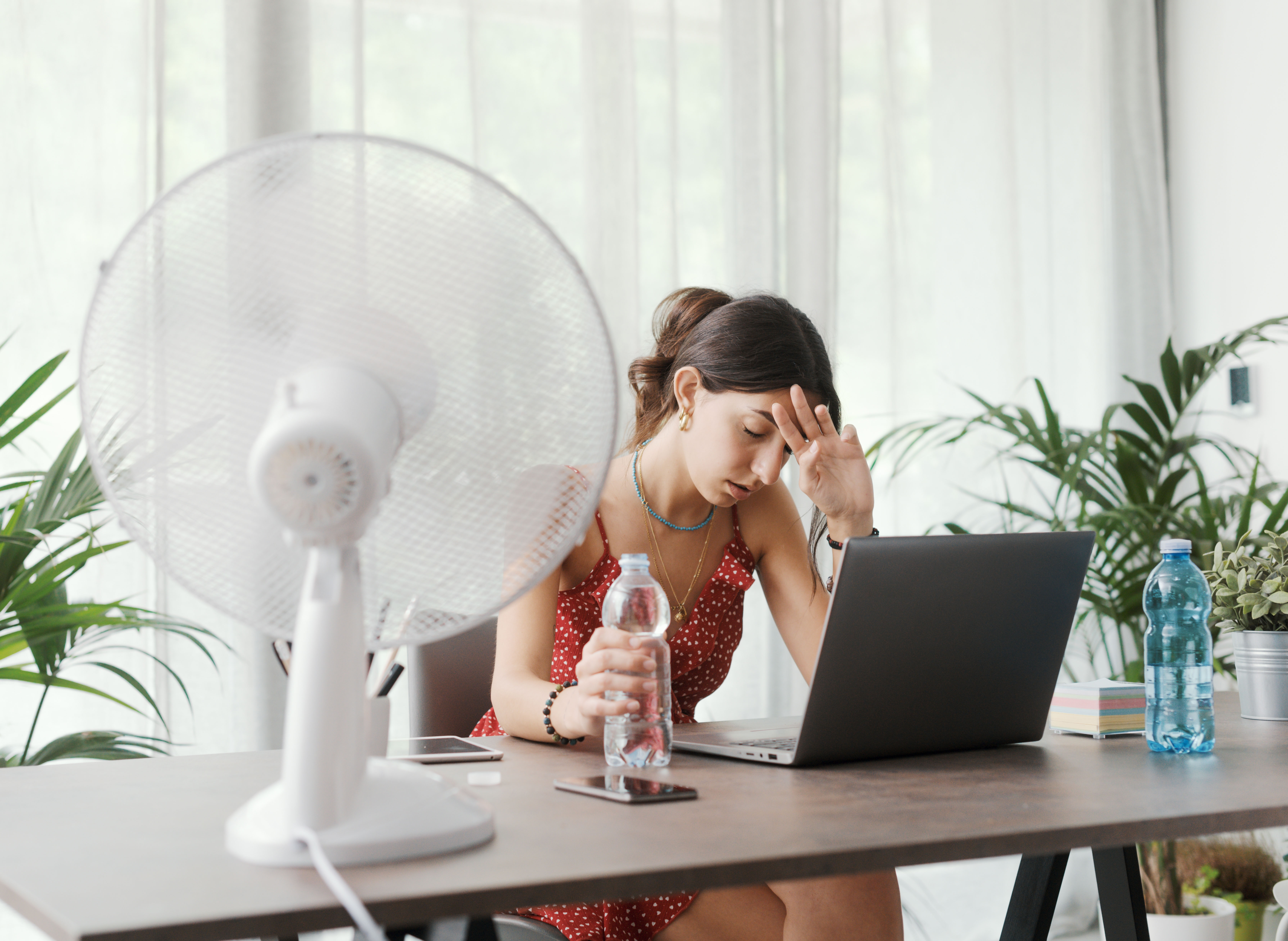 Woman sitting at a desk looking hot with a bottle of water and a desk fan 