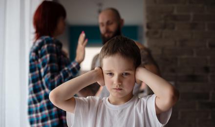 A young child puts his hands over his ears. A woman and man are arguing behind him.