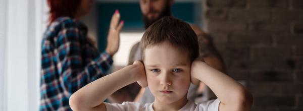 A young child puts his hands over his ears. A woman and man are arguing behind him.