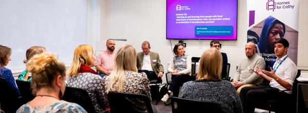 People sitting down in rows listening to six people talk at the front of the room