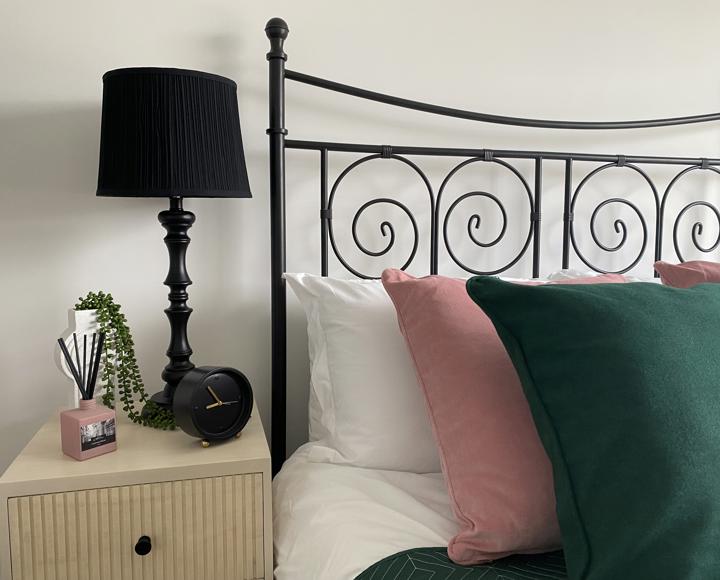 A black wrought iron double bed with green and pink cushions and bedside table