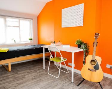 Bedroom with orange wall, a bed, table and chair and guitar