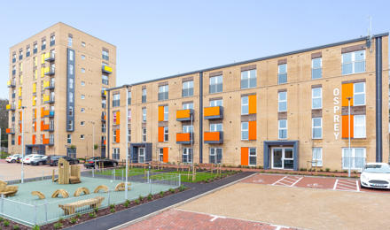 A long row of flats with orange and yellow balconies. In front of them there is a play park.