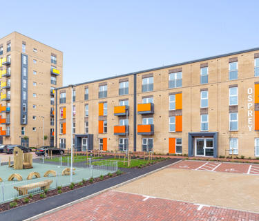 A long row of flats with orange and yellow balconies. In front of them there is a play park.