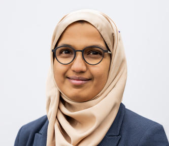 Headshot of a member of Hightown's Board, Zeena Farook. She is young and is wearing glasses and a blue checked blazer.