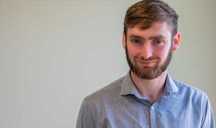 A young male graduate trainee. He is wearing a light grey shirt and has brown hair and facial hair, with blue eyes. 