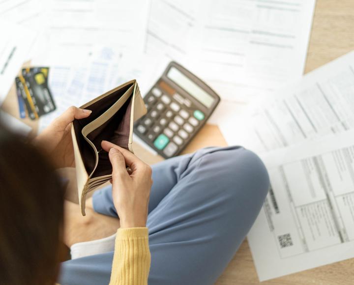 A person is looking inside an empty wallet and has a calculator and paperwork on the floor. They're trying to work out their finances to pay the rent. 