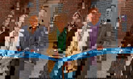 Debbie Jinks, Cllr Mione Goldspink and Andrew Royall. Cllr Goldspink is cutting a ribbon with gold scissors in front of a block of flats.