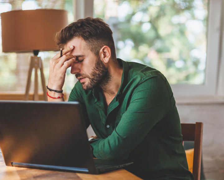 A man looking stressed, sat at a kitchen table. He has a laptop in front of him. 
