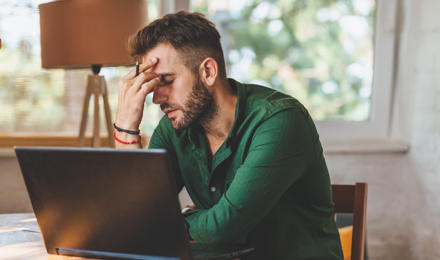 A man looking stressed, sat at a kitchen table. He has a laptop in front of him.