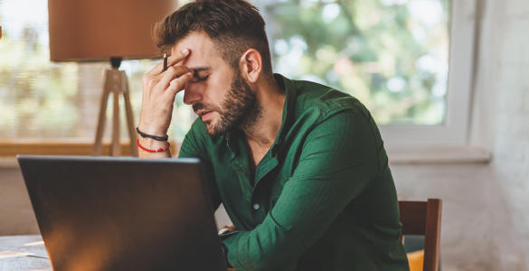 A man looking stressed, sat at a kitchen table. He has a laptop in front of him.