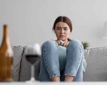 A young woman, sat on the sofa, looking at a bottle and a glass of red wine. She is frowning and looking concerned. 