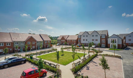 A square communal grass area surrounded by new build flats and houses. Taken on a sunny day. 
