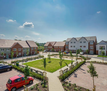 A square communal grass area surrounded by new build flats and houses. Taken on a sunny day. 