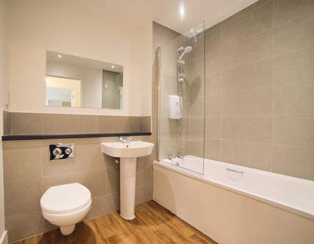 There is a white bathroom suite with a built-in shelf behind the sink and toilet. There is a glass shower screen and shower over the bath.