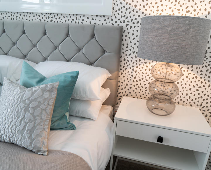 A stock image showing some cushions on a bed with a bedside table and lamp and white wallpaper with black spots on