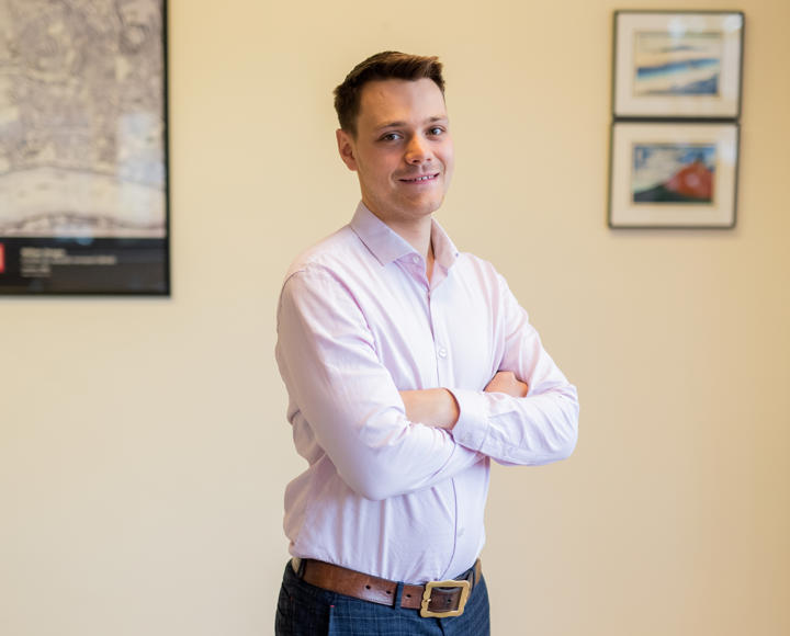 A young male graduate trainee standing up in an office with his arms folded. He is wearing a light pink shirt, a brown belt and navy checked trousers. He has dark brown hair and brown eyes.
