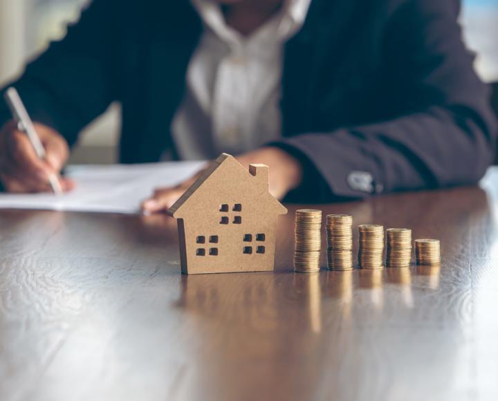 Someone filling in a form and looking at piles of coins, next to the piles of coins there is a small model of a house. 