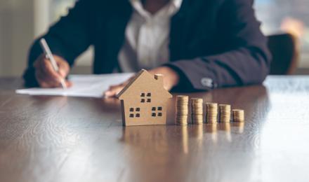 Someone filling in a form and looking at piles of coins, next to the piles of coins there is a small model of a house. 