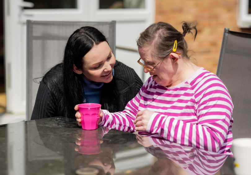 A support worker sitting outside in the sun with a service user, talking and smiling.