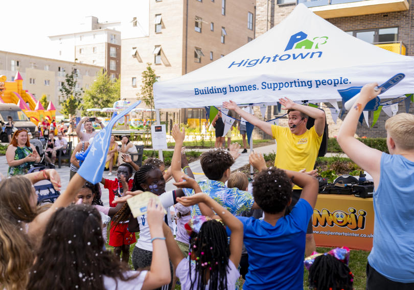 The entertainment at the event waving and getting children to do the same. They are in the sun, there are a lot of children in the photo, and a bouncy castle in the background