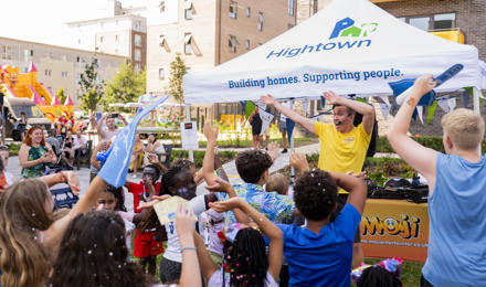 The entertainment at the event waving and getting children to do the same. They are in the sun, there are a lot of children in the photo, and a bouncy castle in the background
