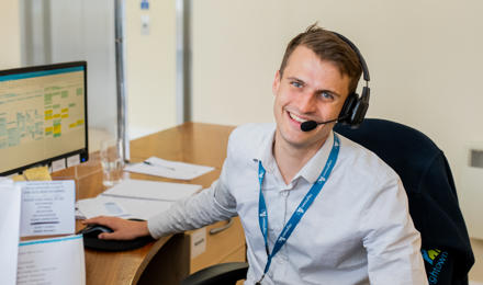 A young male member of staff working on the reception desk. He is wearing a light grey shirt and a headset to take calls.