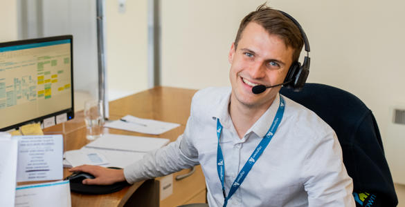 A young male member of staff working on the reception desk. He is wearing a light grey shirt and a headset to take calls.