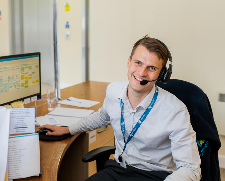 A young male member of staff working on the reception desk. He is wearing a light grey shirt and a headset to take calls.