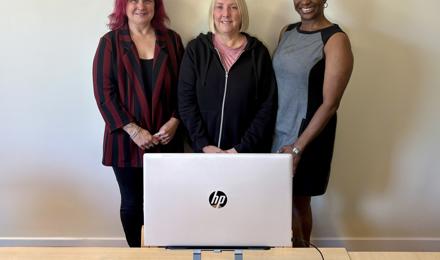 Thre women in the Repairs team smiling standing behind a laptop
