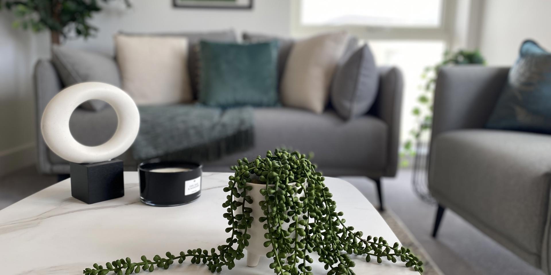 A marble coffee table with a false fern on top. There are grey sofas blurred in the background