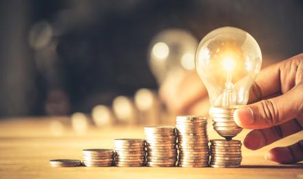 A light bulb is shown next to a small pile of money that is next to a larger pile of money to indicate a saving of energy cost.