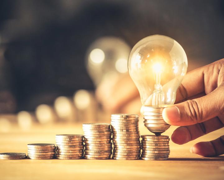 A light bulb is shown next to a small pile of money that is next to a larger pile of money to indicate a saving of energy cost.