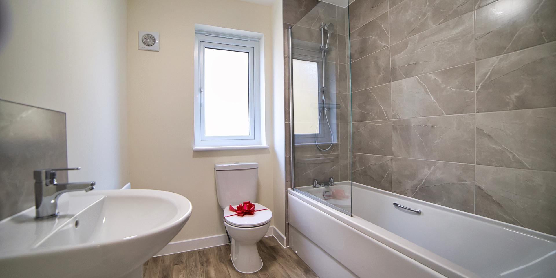 A view of the bathroom with its white suite, shower over the bath, wash hand basin and heated towel rail
