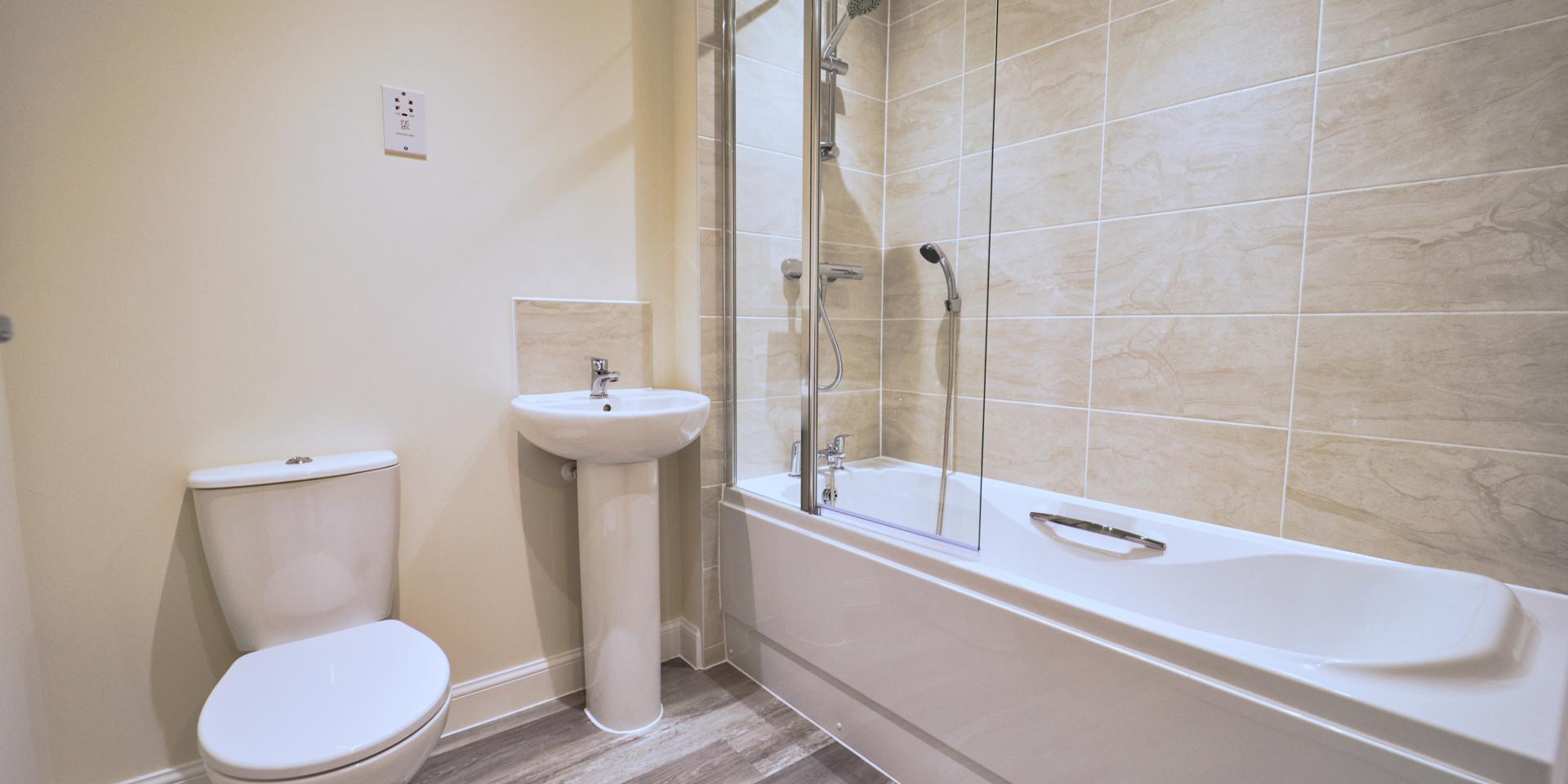 White bathroom suite with shower over bath and shower head attached to taps. Glass shower screen, pedestal sink and toilet