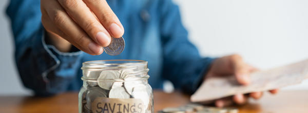 Someone counting coins into a glass jar. The jar is labelled 'savings'. 