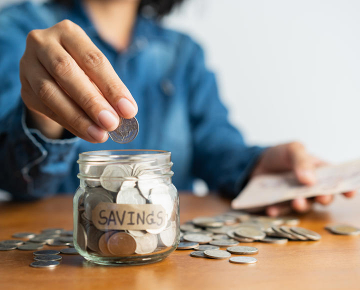 Someone counting coins into a glass jar. The jar is labelled 'savings'. 
