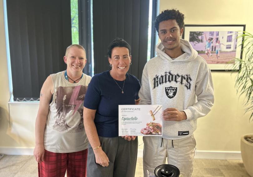 Two hightown staff members and a participant of the cook off smile at the camera and hold up a certificate of participation in front of a table of food at Hightown House