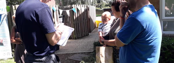 A member of staff is at a summer event, talking to a resident and taking notes outside