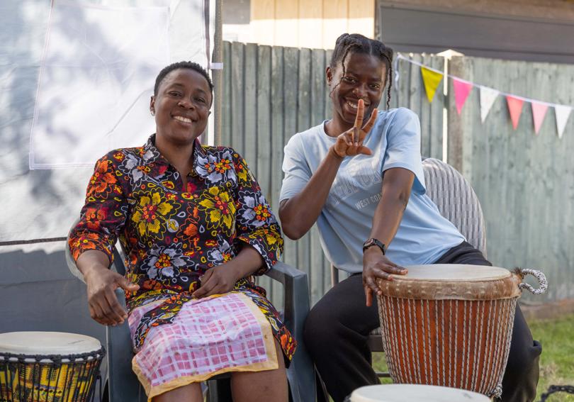 Two people smiling in the sun, while one shows a peace sign and bangs a bongo drum
