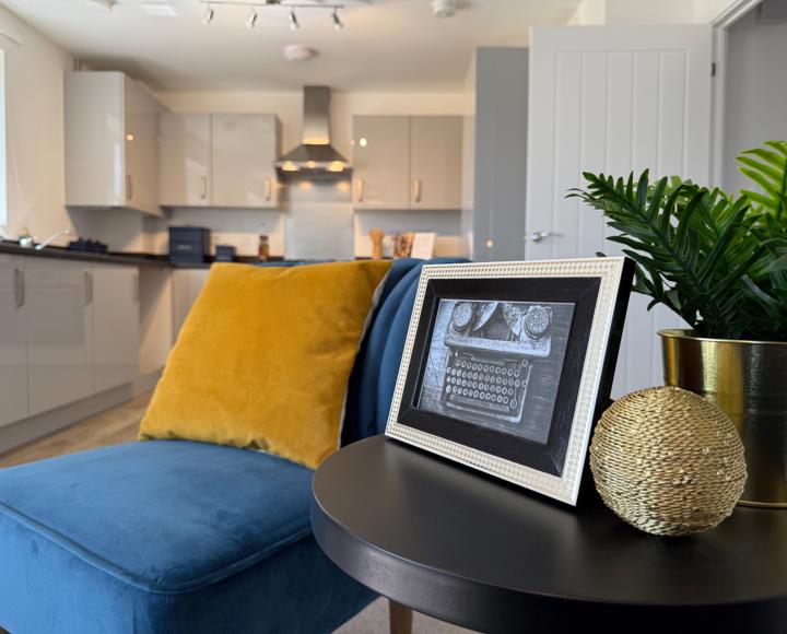 A photo of an old fashioned type writer in a frame, on a side table, next to a blue velvet chair and ochre coloured velvet cushion
