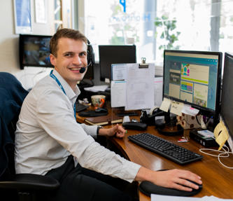 A young male graduate trainee manning the reception desk. He is wearing a light grey shirt and a headset for taking calls. 