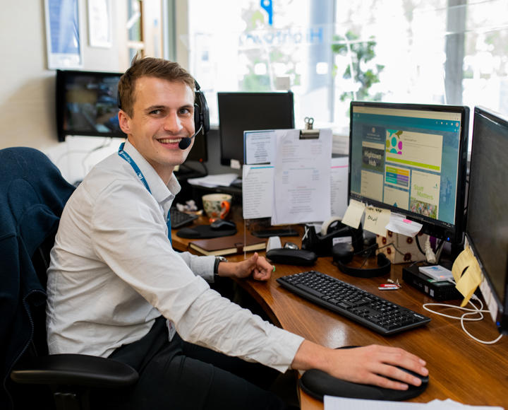 A young male graduate trainee manning the reception desk. He is wearing a light grey shirt and a headset for taking calls. 