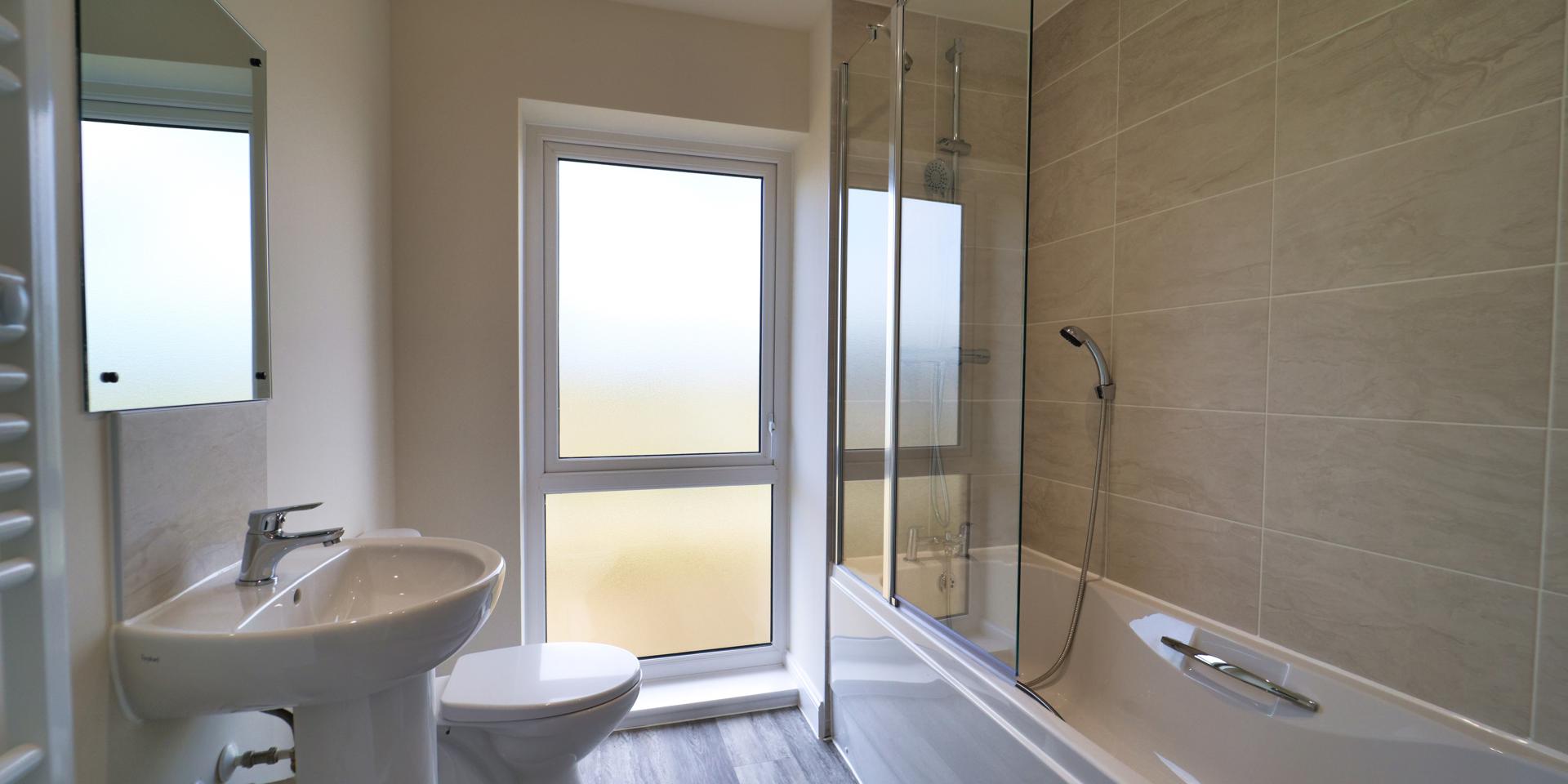 There's a white bathroom suite with a shower over the bath and another shower head attached to the bath taps. There is a large frosted window which goes from the floor to almost the ceiling.