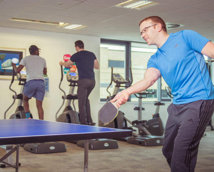 Hightown staff using the gym. One man is playing table tennis and two men are talking whilst on a cross trainer machine.