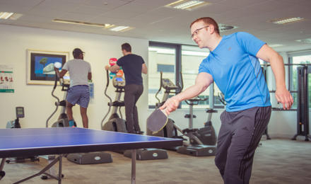 Hightown staff using the gym. One man is playing table tennis and two men are talking whilst on a cross trainer machine.
