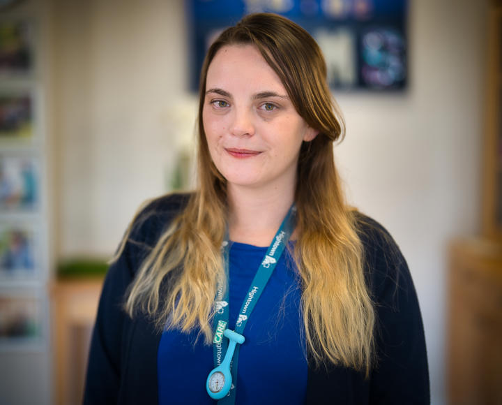 A young female senior support worker looking at the camera. She is wearing a cornflower blue top and a black cardigan.