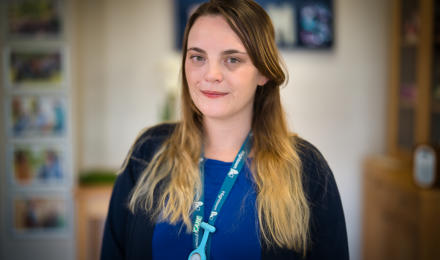 A young female senior support worker looking at the camera. She is wearing a cornflower blue top and a black cardigan.