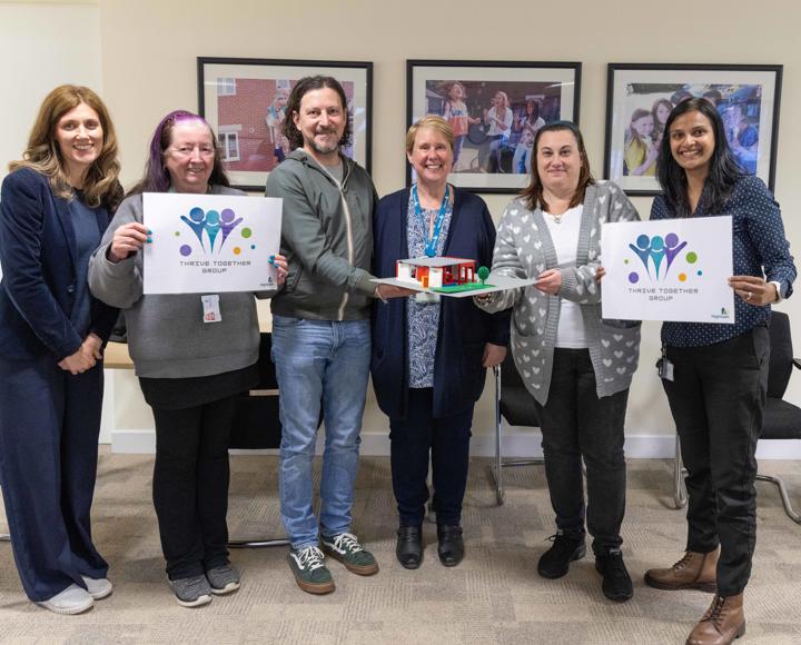 Five women and one man standing up and smiling, holding signs that read Thrive Together Group