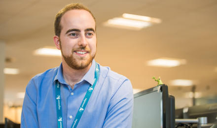 Staff member sat down, looking at the camera, smiling. He is wearing a light blue shirt. 